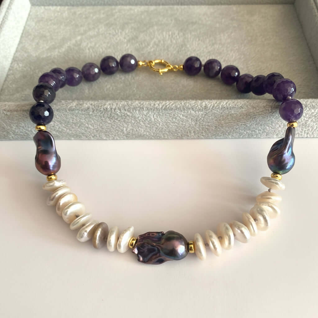 A beaded necklace featuring dark purple amethyst beads, white baroque shell beads, a central iridescent purple bead, and gold spacer rings, displayed on a light gray velvet jewelry box.