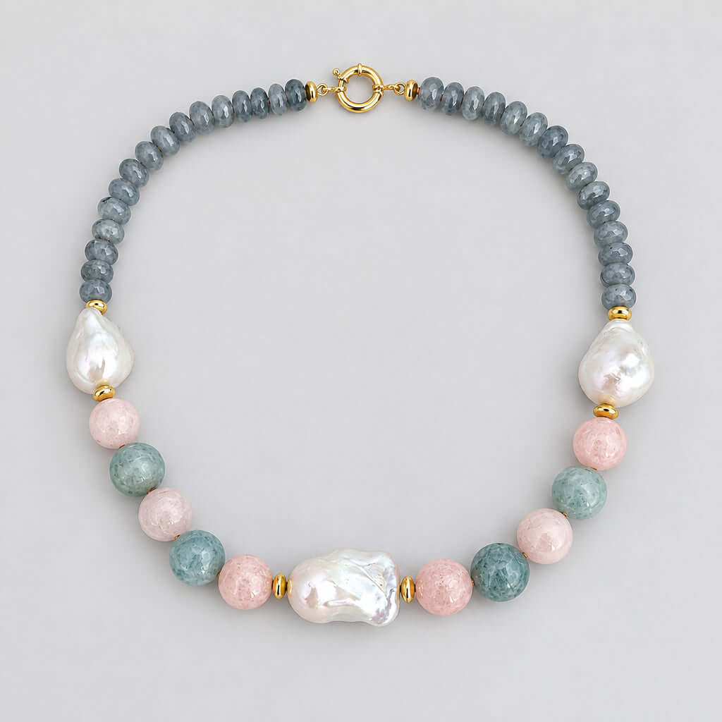 A beaded necklace with a strand of dusty blue smooth beads, accented by white baroque pearls, light pink round beads, light blue-green round beads, and small gold spacer beads, finished with a gold clasp, shown on a plain off-white background.