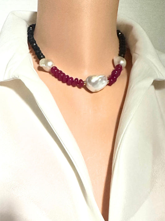 A close-up of a necklace worn on a mannequin torso dressed in a white collared shirt. The necklace features dark black beads, deep red beads, white pearl-like round beads, and a large irregular white baroque pearl at the center.