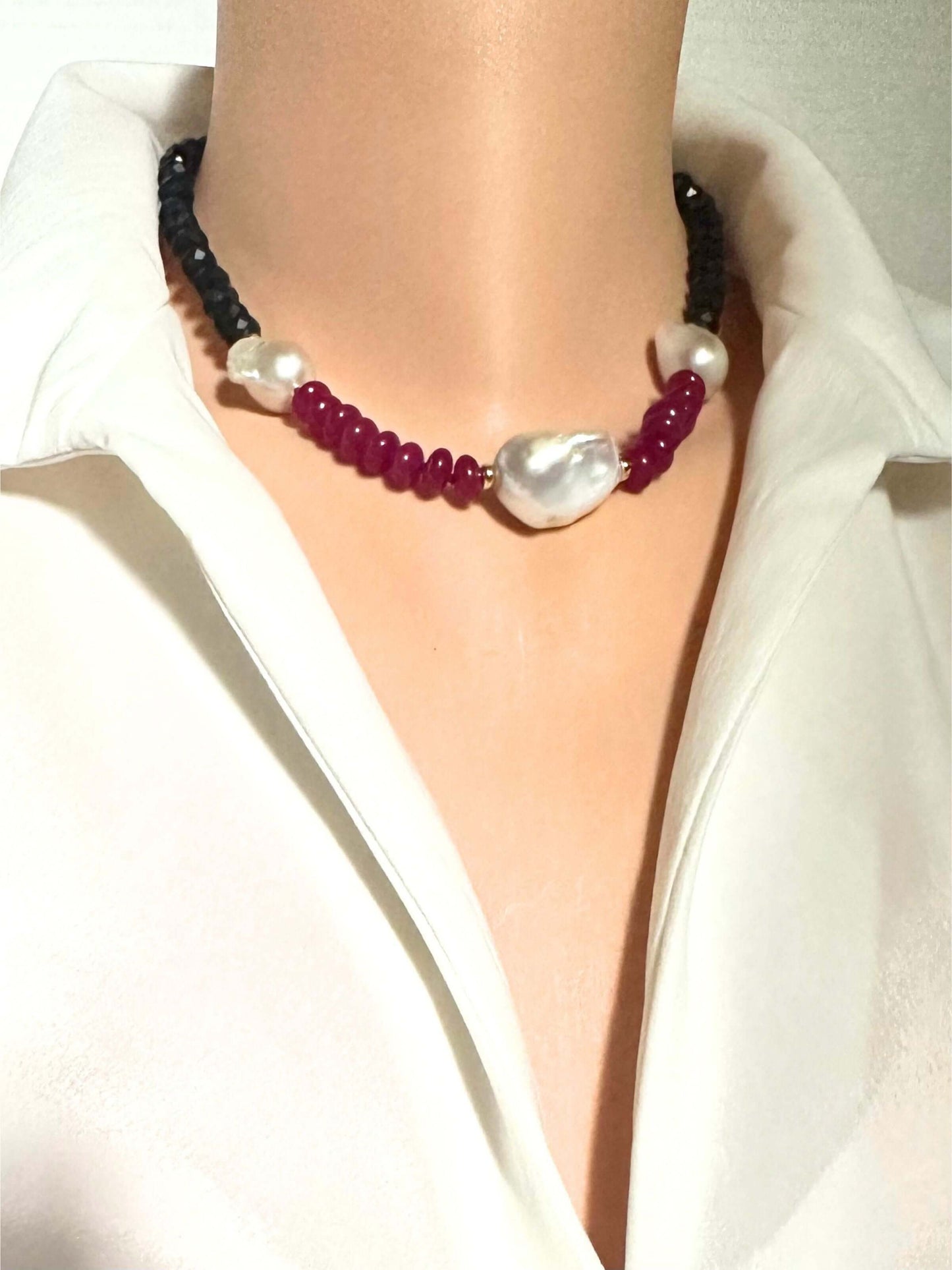 A close-up of a necklace worn on a mannequin torso dressed in a white collared shirt. The necklace features dark black beads, deep red beads, white pearl-like round beads, and a large irregular white baroque pearl at the center.