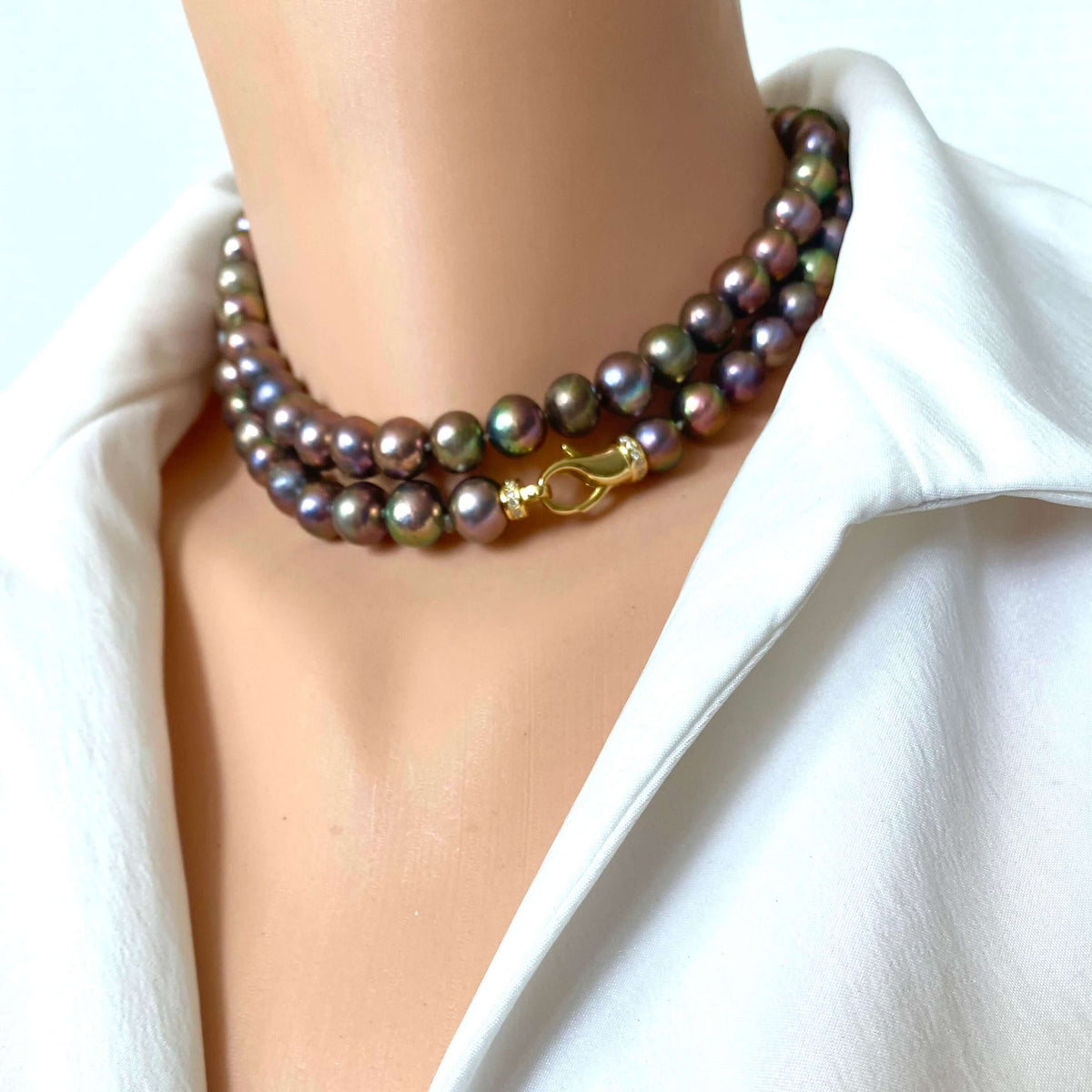A close-up of a double-strand pearl necklace with iridescent dark gray-green beads and a gold clasp, worn on a mannequin neck over a white collared shirt.