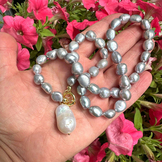 Grey pearl necklace with white baroque pearl pendant displayed in hand among colorful flowers.