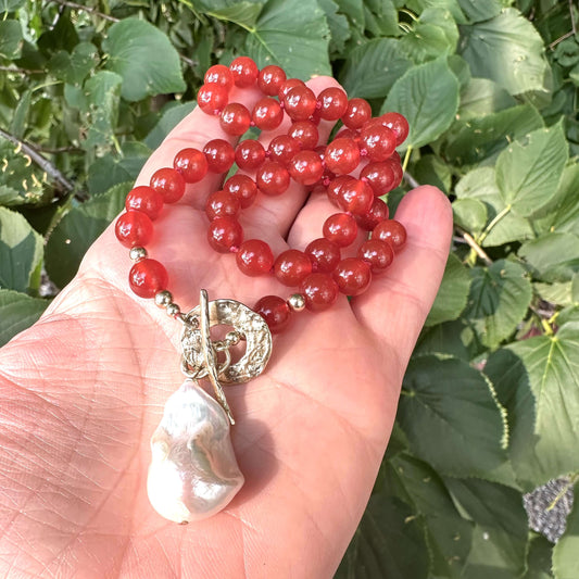 Red agate toggle necklace with baroque pearl pendant held in hand against green foliage