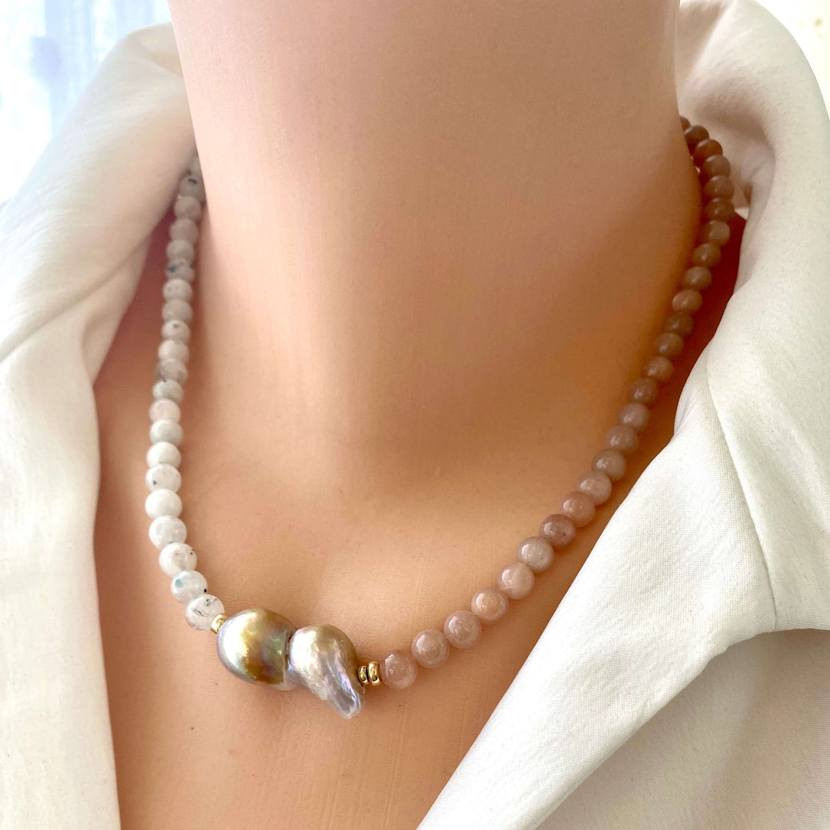 A close-up of a pearl necklace displayed on a mannequin neck wearing a white collared shirt. The necklace features small, round pale pinkish-beige pearls, a small clear faceted bead, and two larger baroque-style pearls in iridescent gold, silver, and lavender tones.