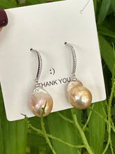 Load image into Gallery viewer, Golden pink baroque pearl dangle earrings with CZ pavé silver hooks held near green leaves, handcrafted in Canada.