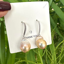 Load image into Gallery viewer, Golden pink baroque pearl dangle earrings displayed against green leaf backdrop, designed and handmade in Canada.