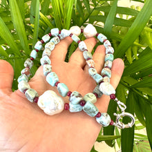 Load image into Gallery viewer, Hand holding larimar ruby and baroque pearl necklace near green leaf background
