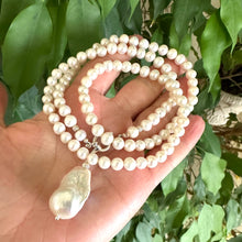 Load image into Gallery viewer, Hand holding white pearl necklace with baroque pendant near green leaf background