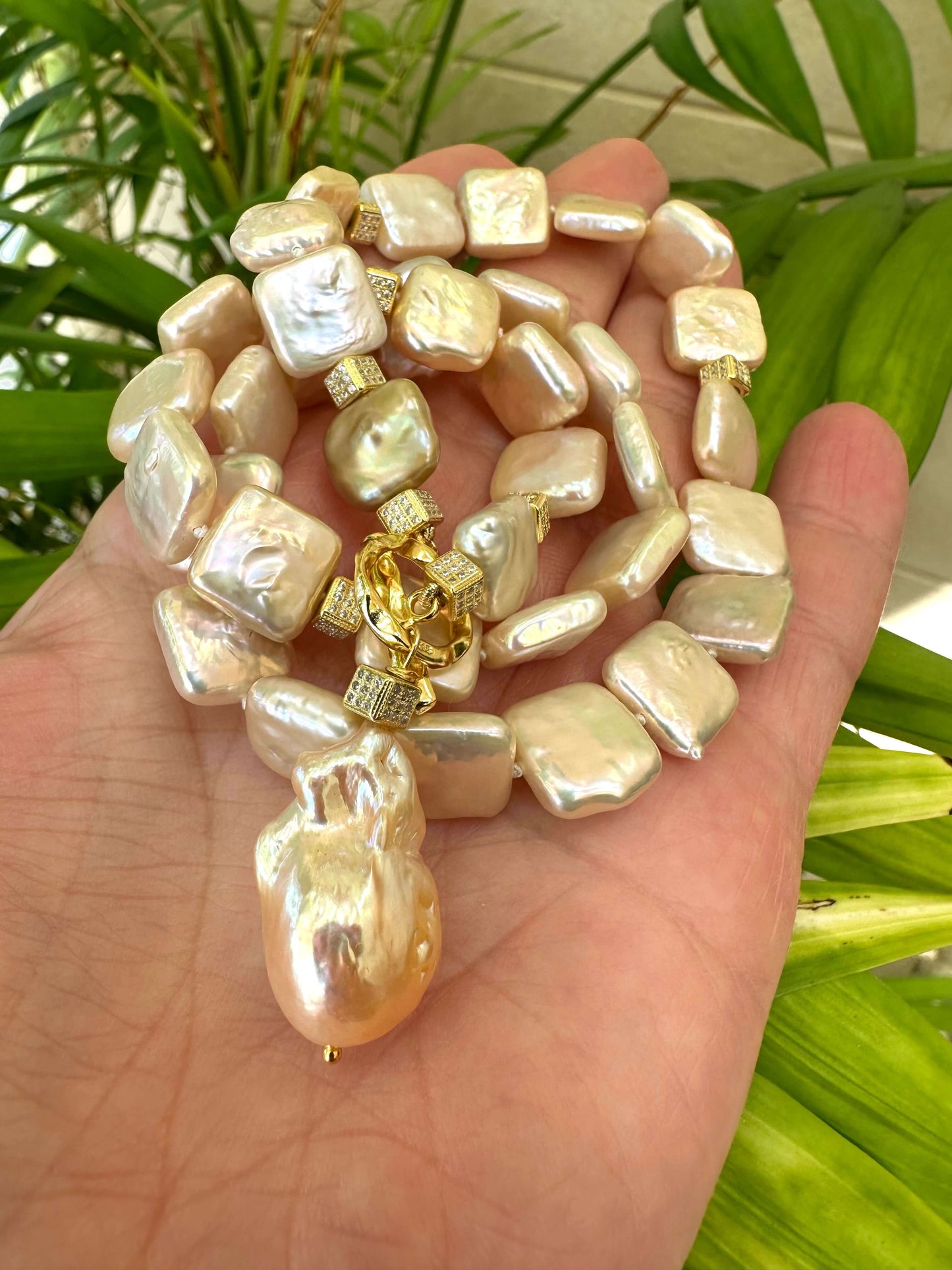 Hand holding golden baroque pearl toggle necklace against leafy green background — lustrous baroque pearls and gold details by Loulia Pearl Jewelry