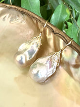 Load image into Gallery viewer, White baroque pearl drop earrings resting on green leaf and pearl shell backdrop, silver hooks visible