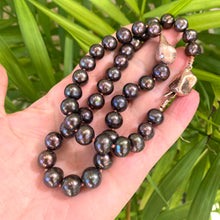 Load image into Gallery viewer, Black pearl necklace with rose gold pendant held in hand against green leaf backdrop