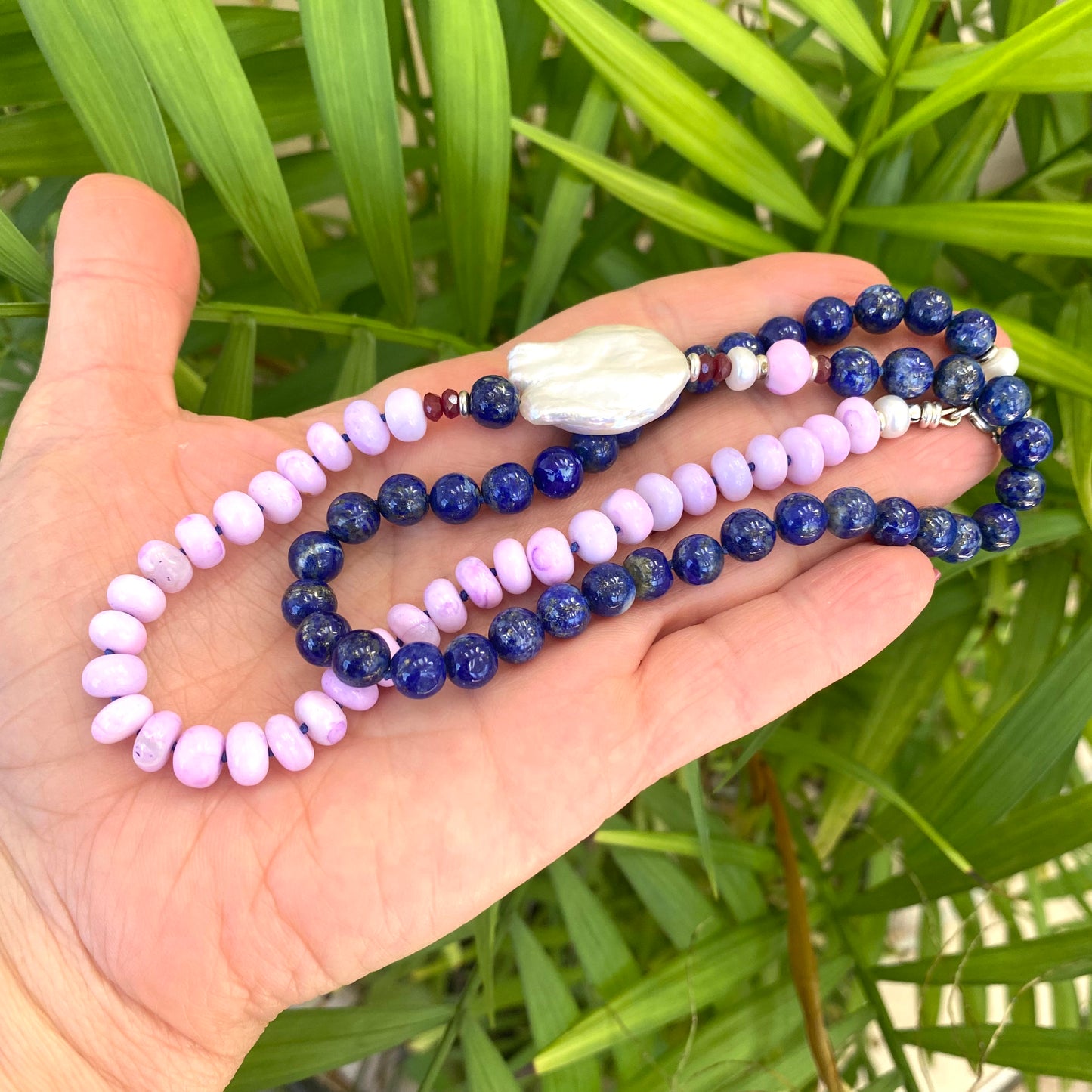 Lapis lazuli and baroque pearl necklace held in hand against green leaves, natural gemstone jewelry.