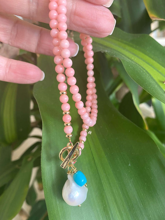 Pink coral toggle necklace with baroque pearl and turquoise charm against green leaves.