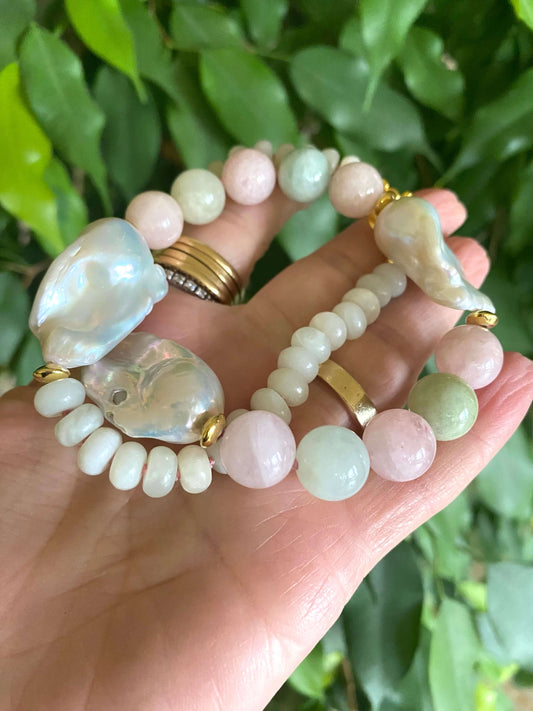 Hand holding morganite and jade necklace with baroque pearls against green natural foliage, showing gemstone color and luster.