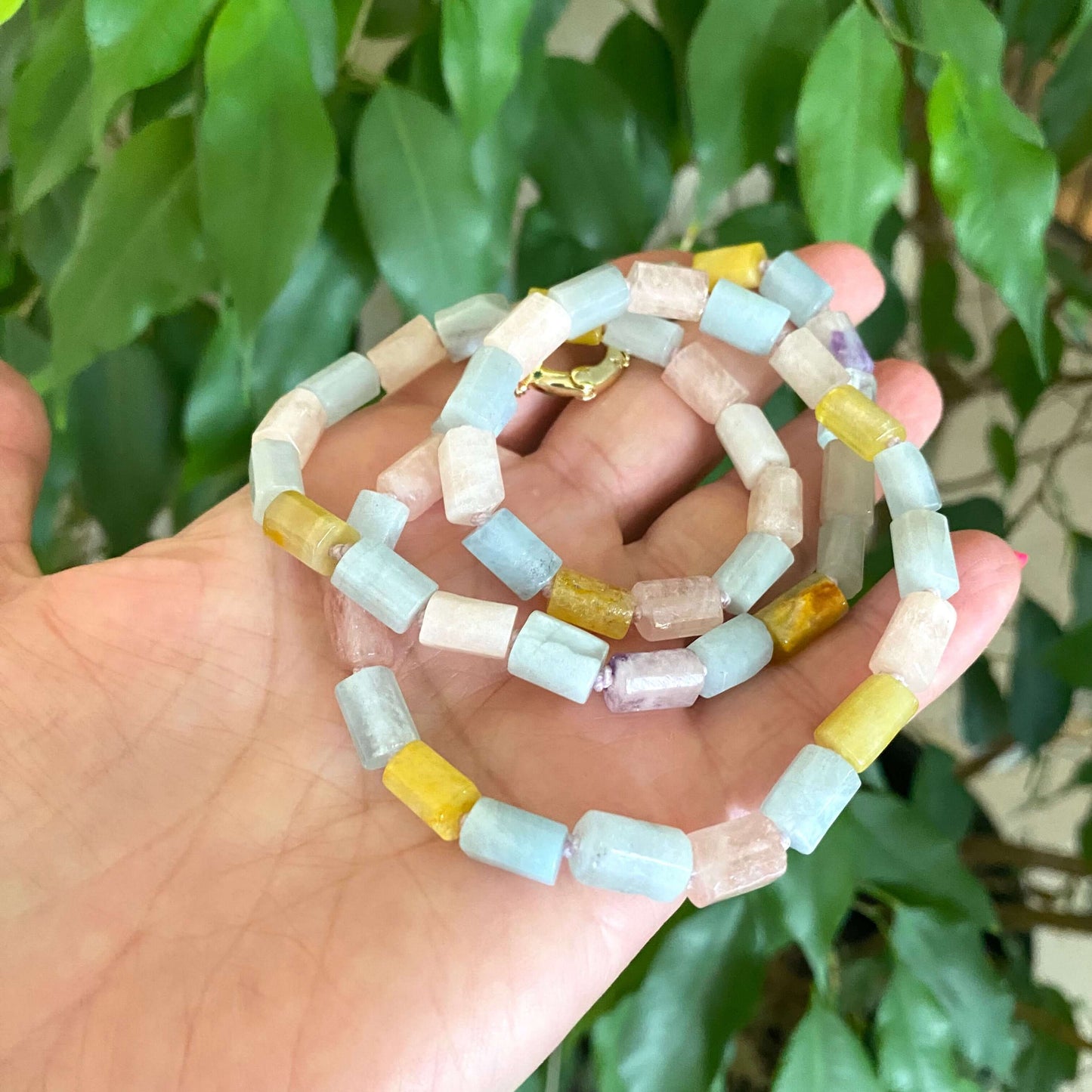 Beryl gemstone necklace featuring aquamarine, morganite, heliodor, and goshenite on a hand against green foliage.