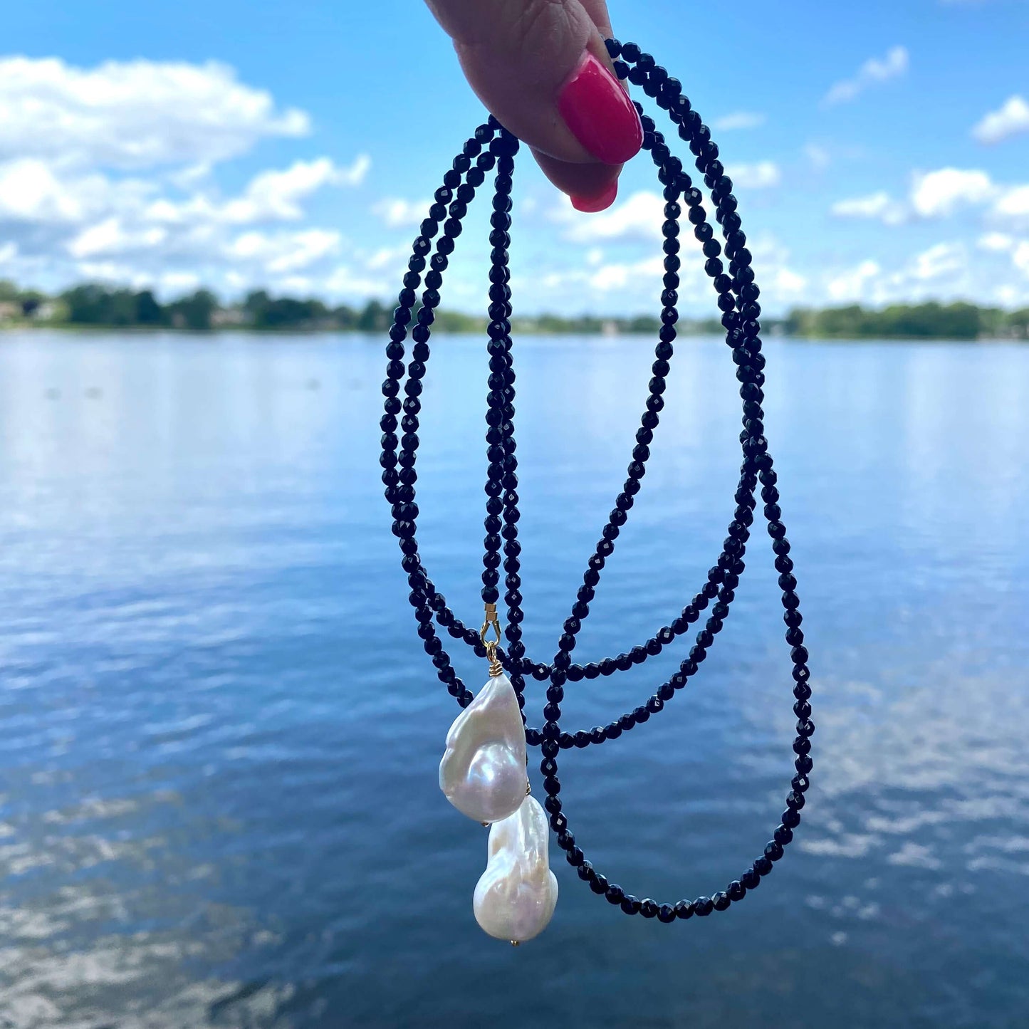 Black spinel lariat necklace with baroque pearl, handmade in Canada, showcased against a scenic lake background.