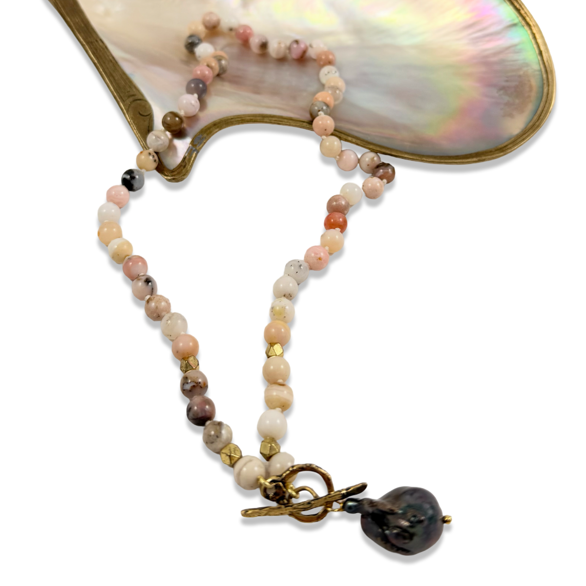 A beaded necklace with polished and faceted beads in soft peach, cream, pink, gray, and brown tones, accented with small gold spacer beads and a large dark brown baroque toggle clasp, displayed on a vintage iridescent shell fan against a white background.