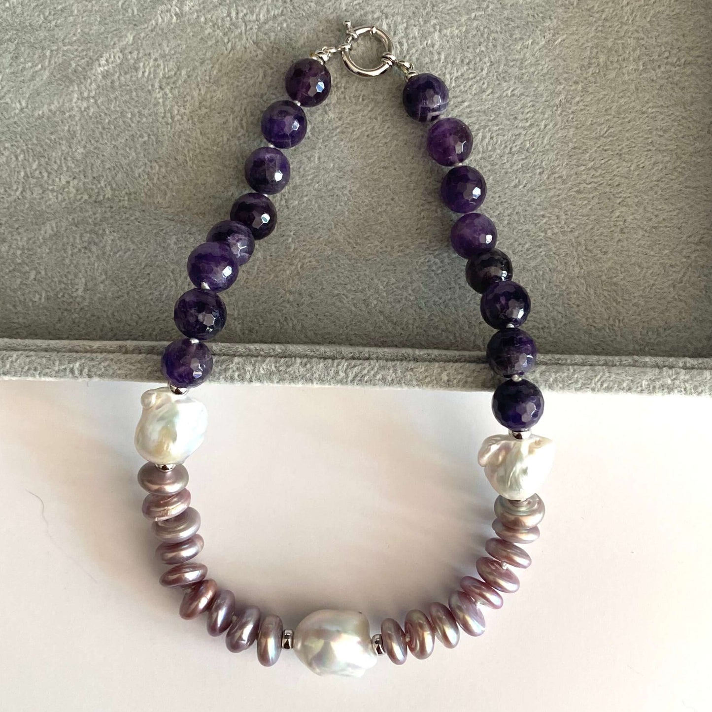 A beaded necklace with dark purple round beads, white pearl-like beads, and metallic bronze disc beads, displayed on a gray velvet jewelry box insert.