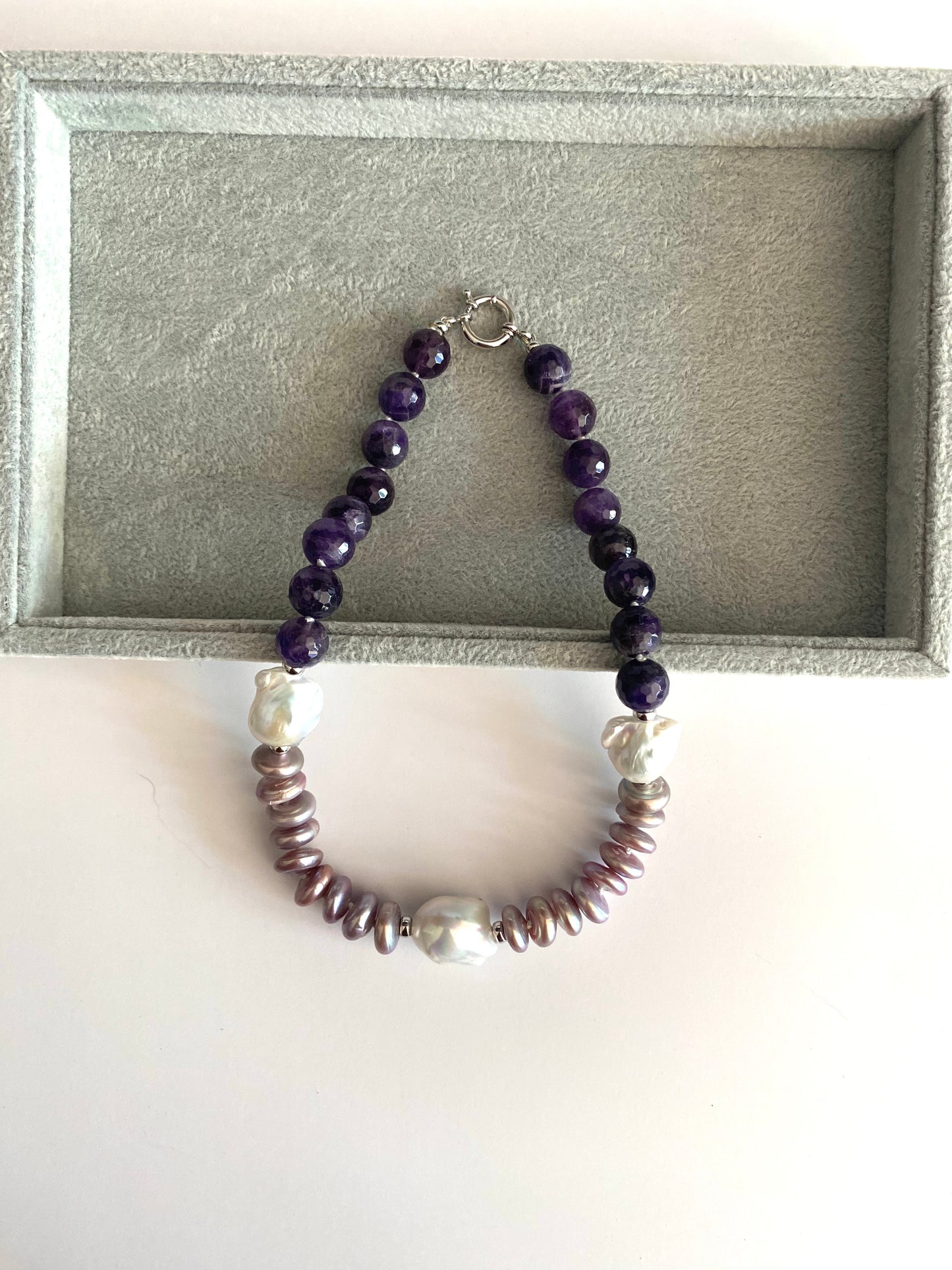 A beaded necklace with dark purple round beads, white pearl-like beads, and metallic bronze disc beads, displayed on a gray velvet jewelry box insert.