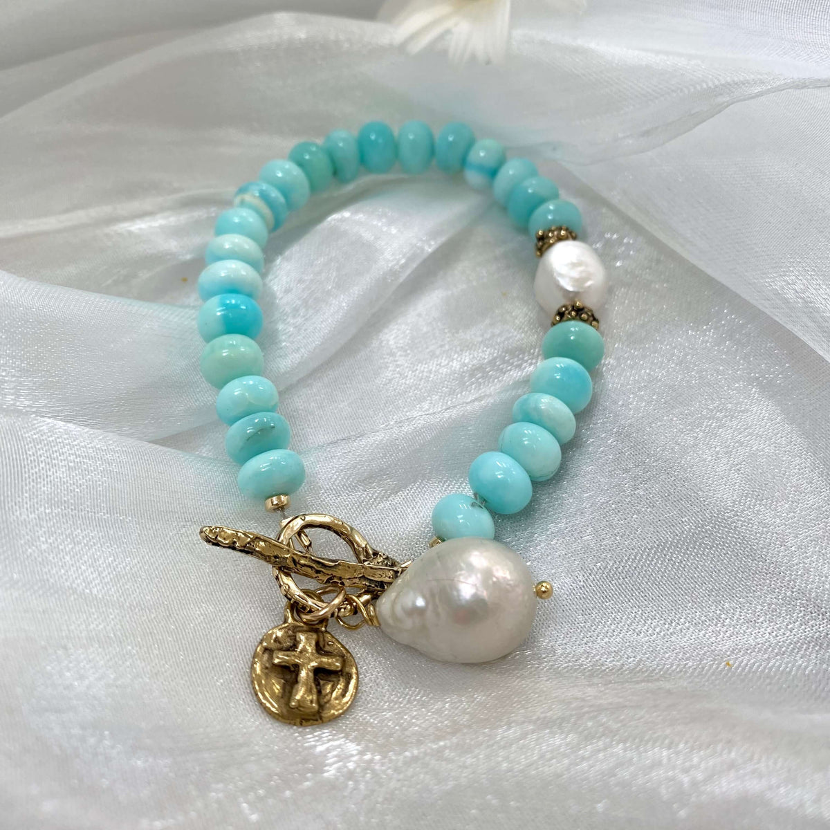 Sky blue opal bracelet with baroque pearls and cross charm on soft fabric background.
