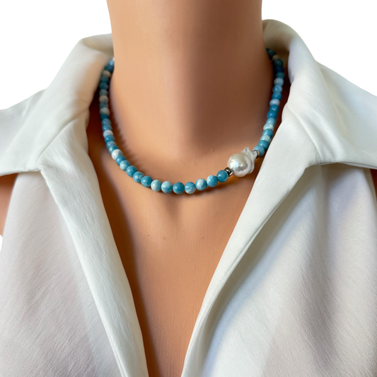 A close-up of a beaded necklace worn on a mannequin or model's neck, featuring light blue round beads and a single white pearl accent, styled with a white collared blouse against a white background.