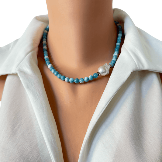 A close-up of a beaded necklace worn on a mannequin or model's neck, featuring light blue round beads and a single white pearl accent, styled with a white collared blouse against a white background.