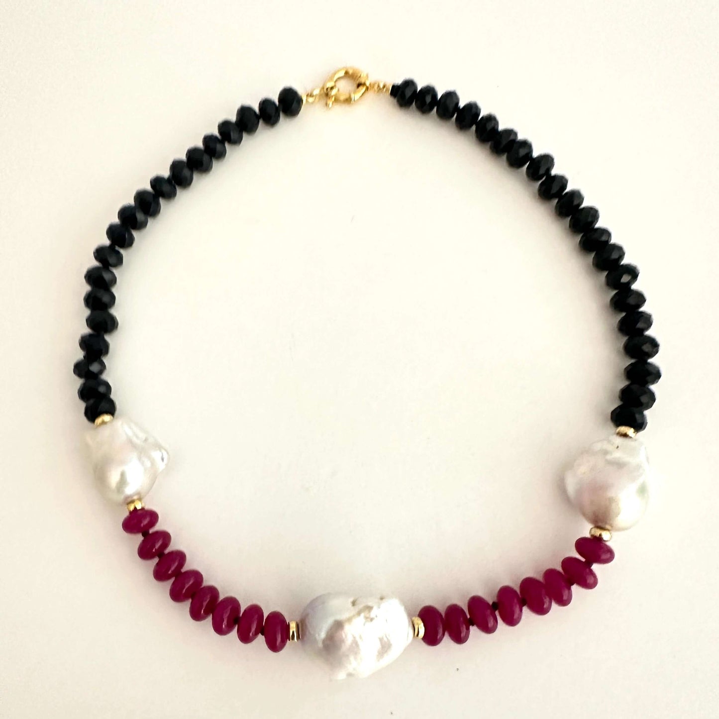 A beaded necklace featuring black faceted beads, deep burgundy round beads, and three white baroque pearls, accented with small gold spacers and a gold clasp, displayed on a light cream background.