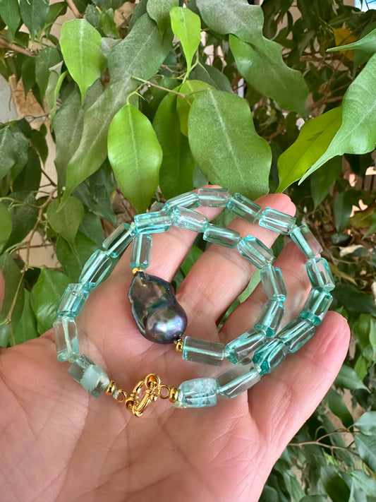 Hand holding a green beaded short necklace with a dark baroque-shaped pendant against a leafy background