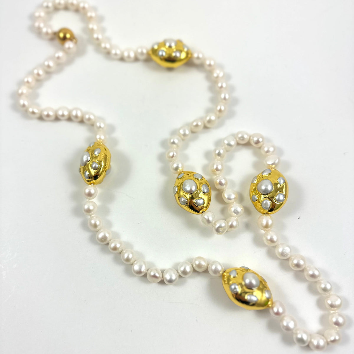 A vintage-style pearl necklace with five ornate gold-toned oval medallions, each decorated with small white pearls, laid out on a plain white surface.
