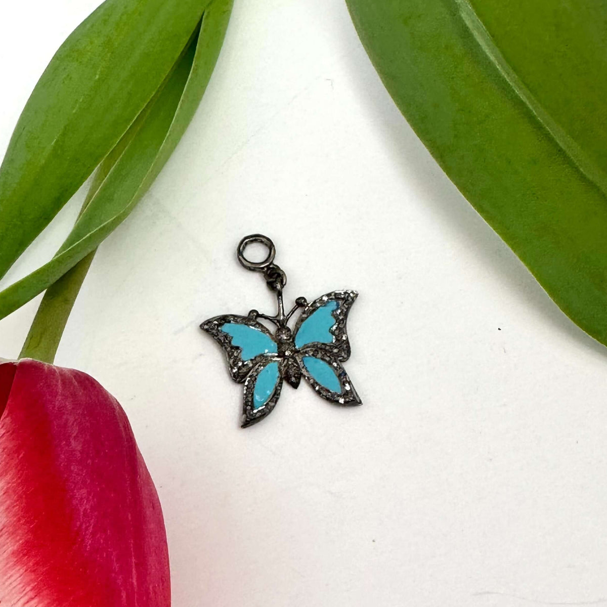 Blue enamel butterfly pendant with diamond pavé displayed next to a red tulip and green leaves.
