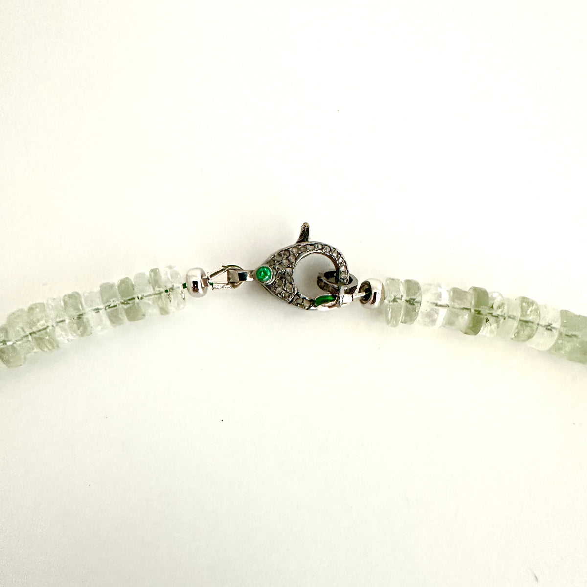 A close-up of a beaded necklace with light green translucent gemstone beads and a silver-toned fish-shaped clasp with a small green stone, shown on a plain white background.
