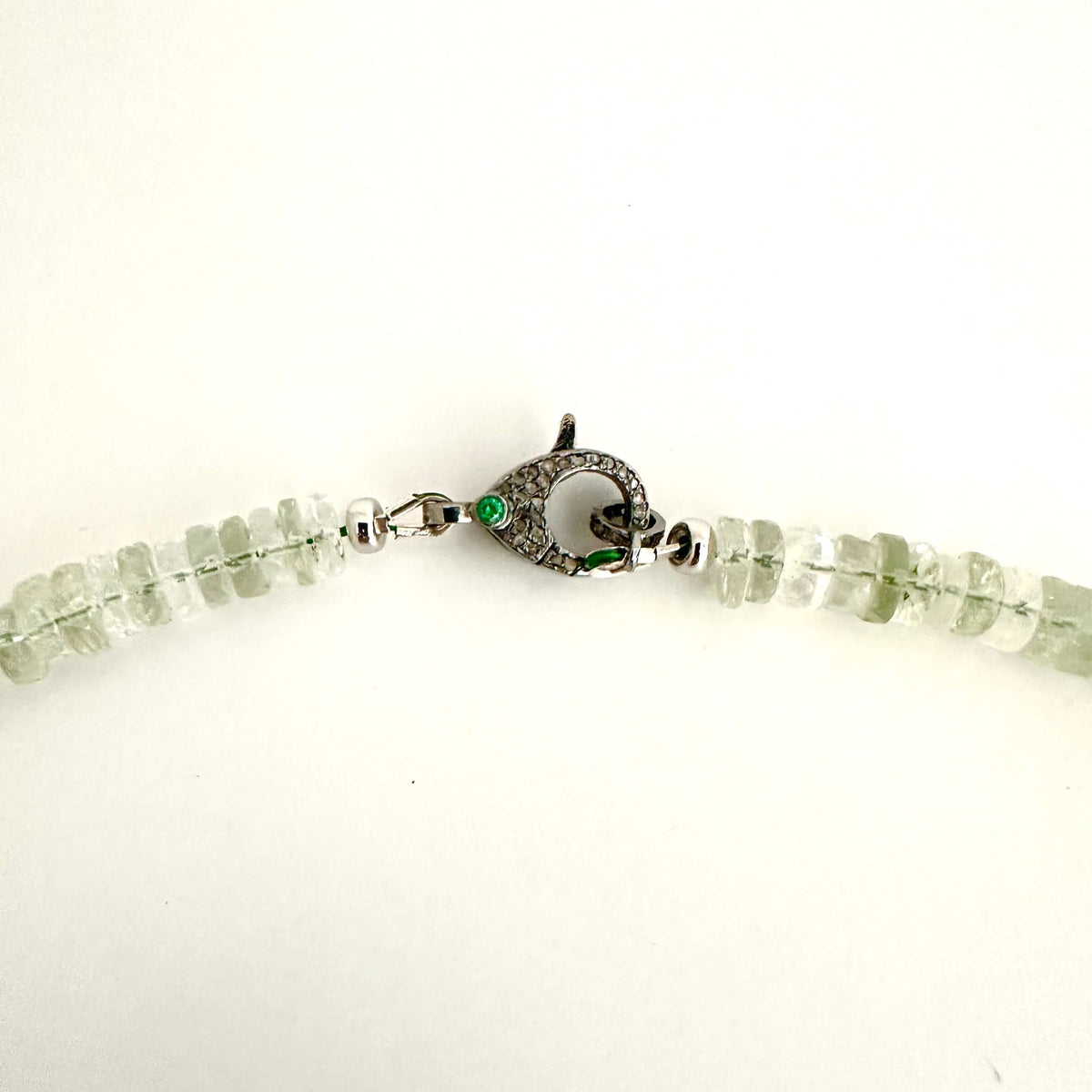 A close-up of a beaded necklace with light green translucent gemstone beads and a silver-toned fish-shaped clasp with a small green stone, shown on a plain white background.