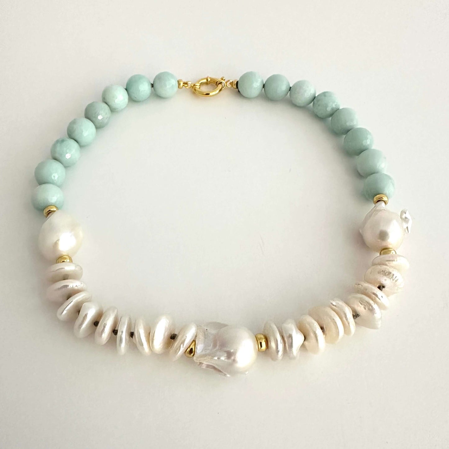 A beaded necklace featuring light mint green round beads, white baroque pearls, and textured white disc beads, accented with small gold spacer rings and a gold clasp, displayed on a light gray background.