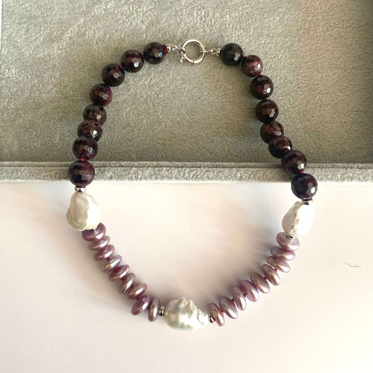 A beaded necklace with dark reddish-brown faceted beads, light purple baroque pearls, and a strand of small lavender beads, displayed on a gray jewelry box.