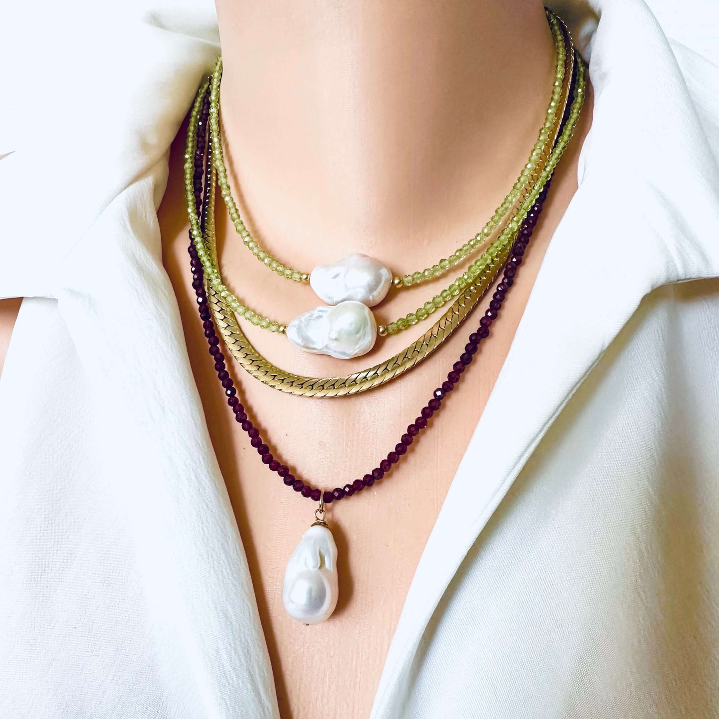 A close-up of a person wearing a layered necklace set with a white baroque pearl pendant, two gold chain necklaces, and dark purple beaded strands, styled with a white collared shirt.