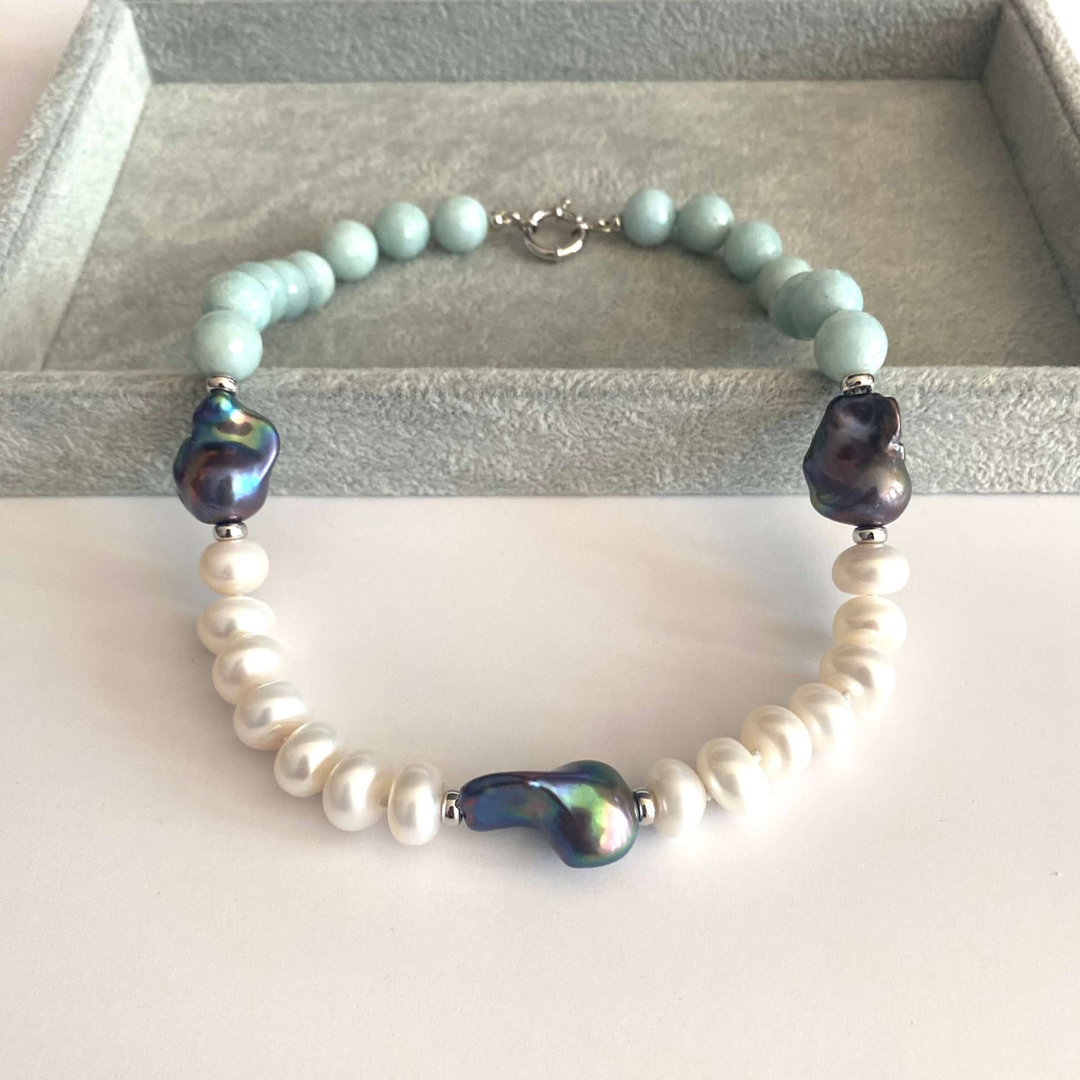 A beaded bracelet displayed on a gray jewelry box. The bracelet features light blue-green round beads, white baroque pearls, and three dark iridescent beads, with a silver clasp.