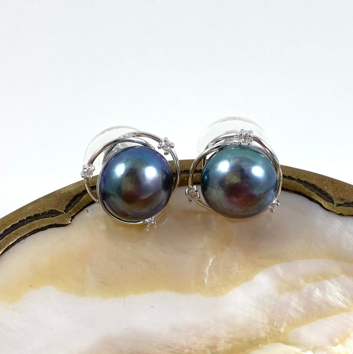 A pair of silver stud earrings featuring round, iridescent dark blue-gray pearls, displayed on a polished shell surface against a white background.
