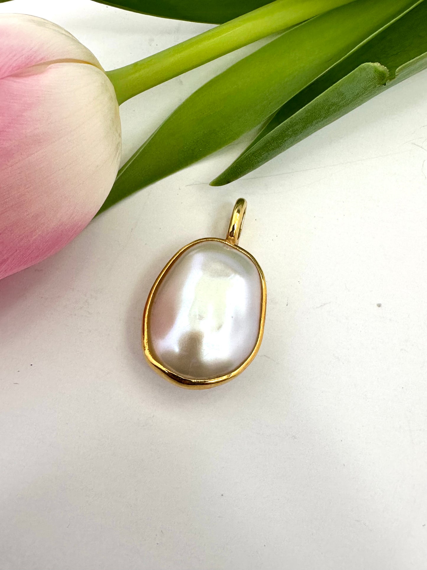 Gold pendant with a large white pearl on a white surface with pink tulip and green leaves.