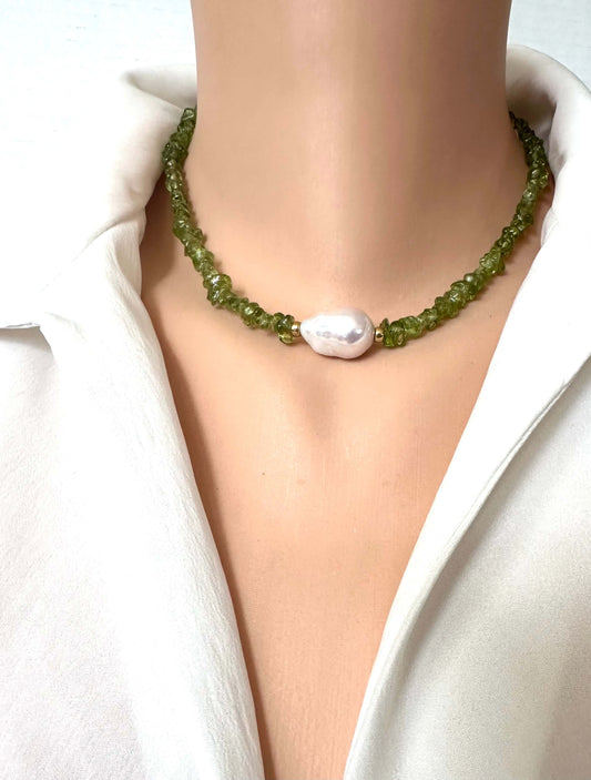 A close-up of a beaded necklace worn on a mannequin or model neck, featuring green faceted beads and a single irregular white pearl at the center, styled with a white collared shirt.