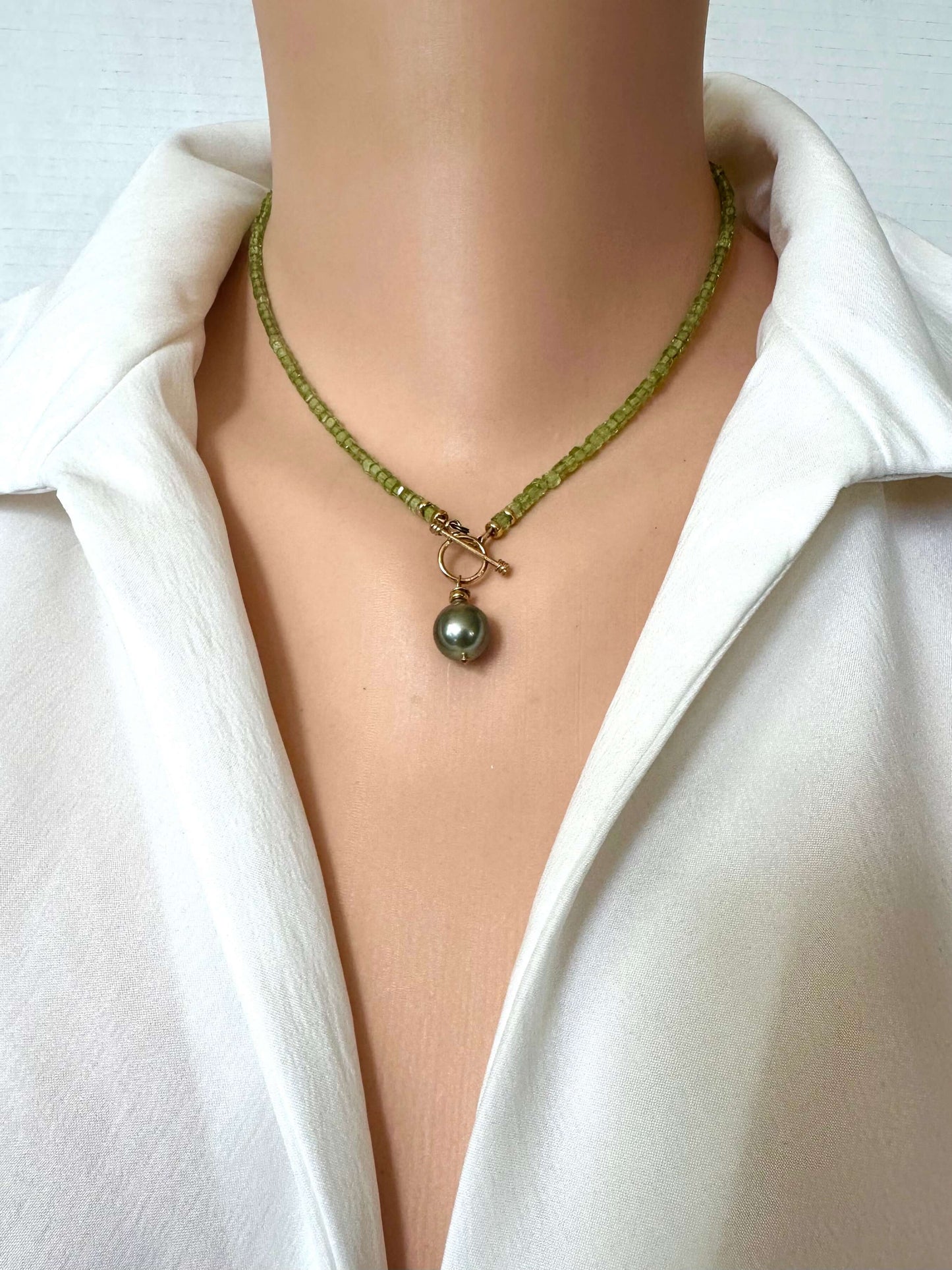A close-up of a necklace worn on a mannequin or model torso, featuring a beaded green chain with a toggle clasp and a dark pearl pendant, styled with a white collared shirt.