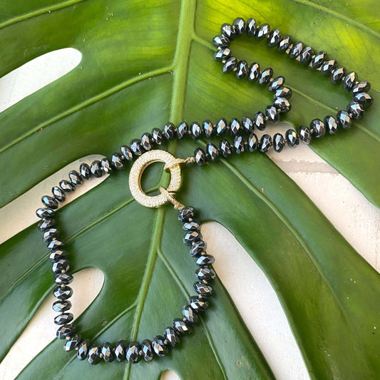 Festive Hematite Necklace featuring faceted hematite beads embraced by a gold statement clasp on a tropical leaf background.