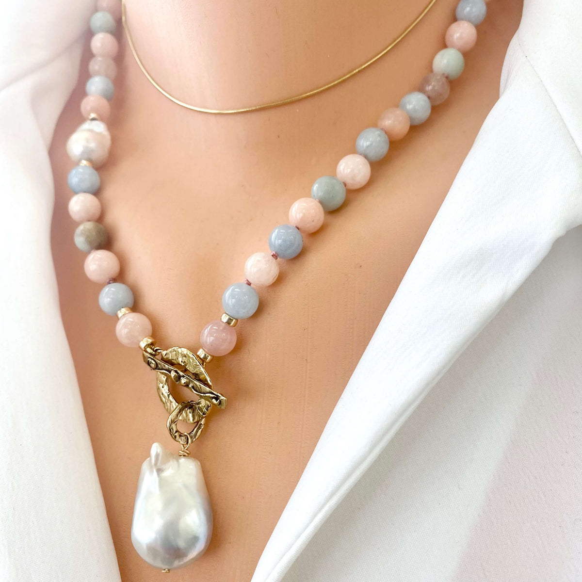 A close-up of a beaded necklace worn on a model's neck, featuring pastel pink, light blue, and gray round beads with a gold clasp and a large white baroque pearl pendant, layered with a thin gold chain.