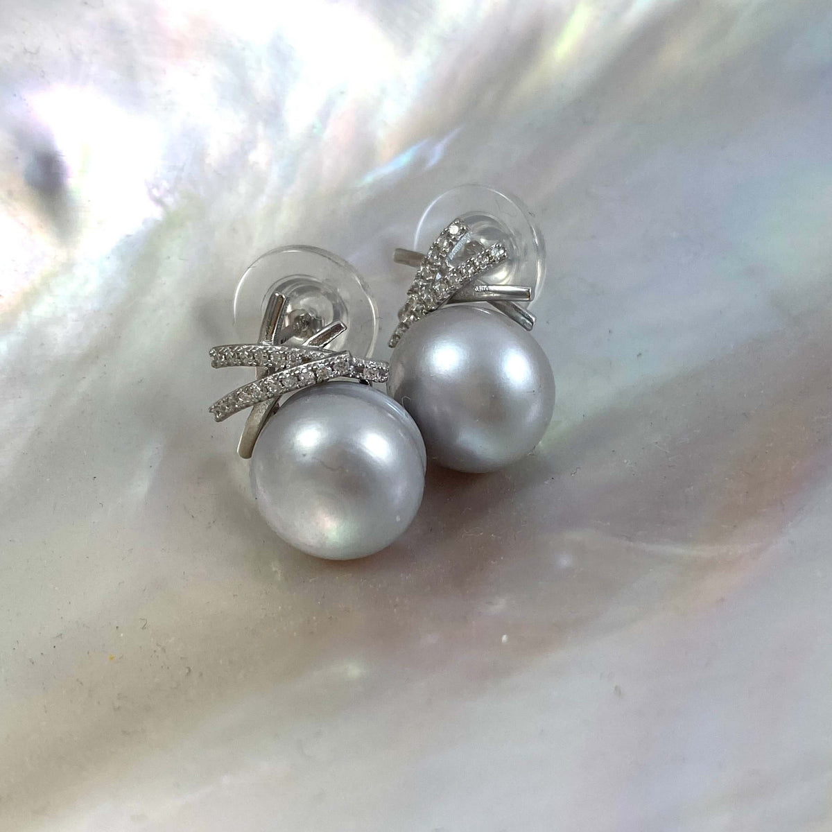 A pair of silver stud earrings with round white pearls and small clear crystal accents, displayed on a shimmering iridescent shell surface.