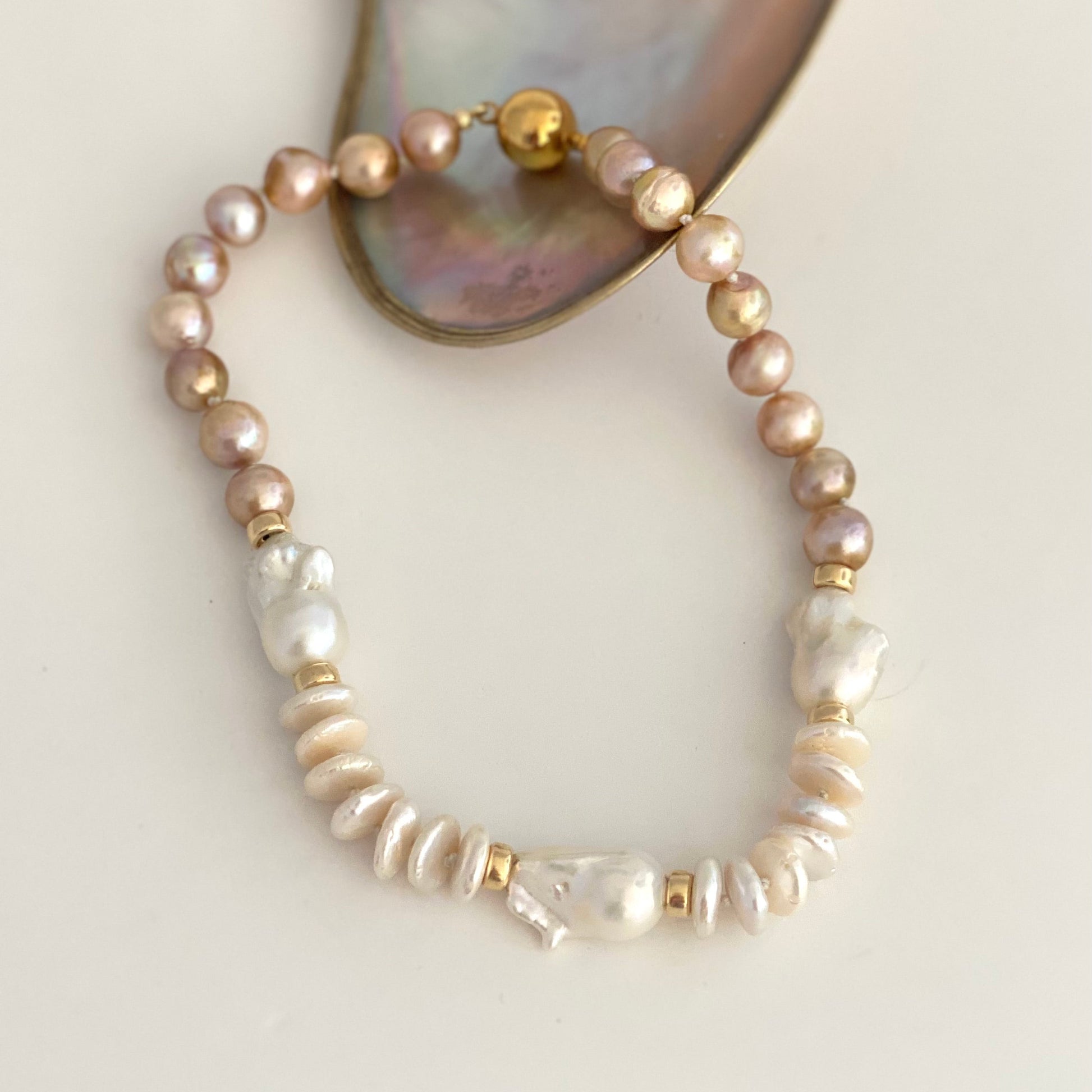 A beaded stretch bracelet featuring light pink freshwater pearls, baroque pearls, and gold spacer beads, displayed on a neutral cream background with a mother-of-pearl shell prop.