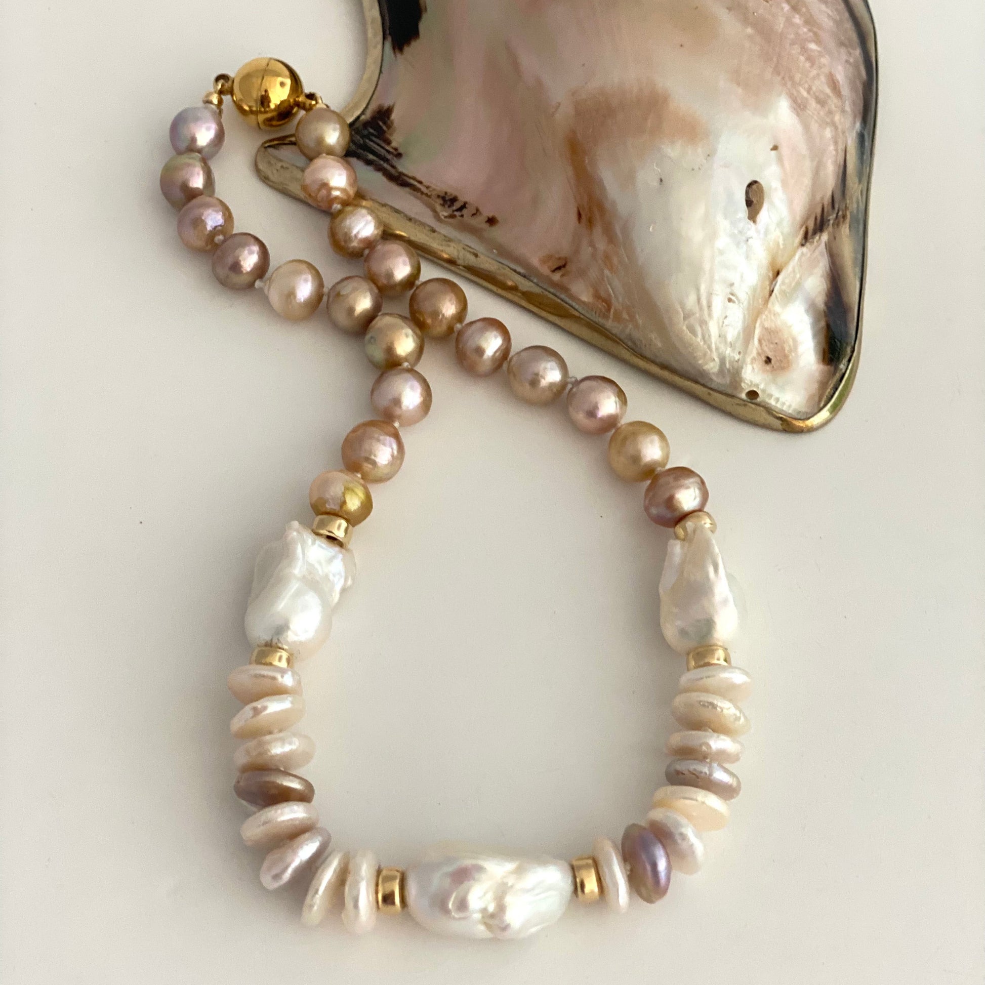 A beaded necklace featuring iridescent baroque pearls in soft pink, beige, and white, accented with textured shell beads and gold spacer rings, displayed next to a large iridescent abalone shell on a light beige background.