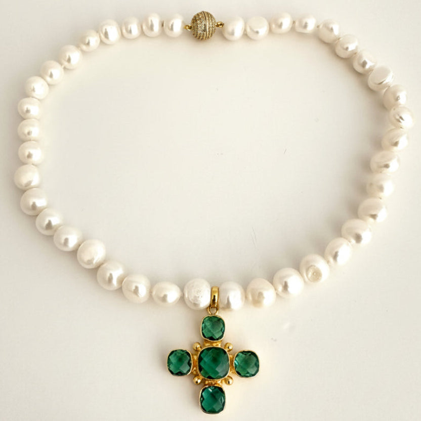 A pearl necklace with a gold cross pendant featuring green gemstones, displayed on a light off-white background.