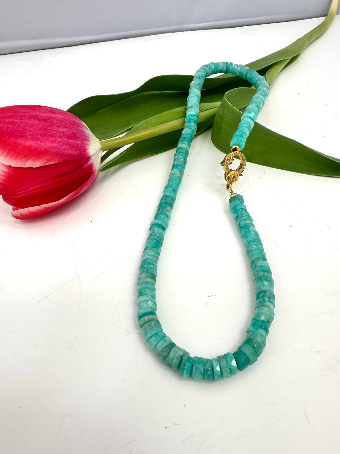 AMAZONITE beaded necklace with a gold clasp on a white surface with a pink tulip and green leaves.