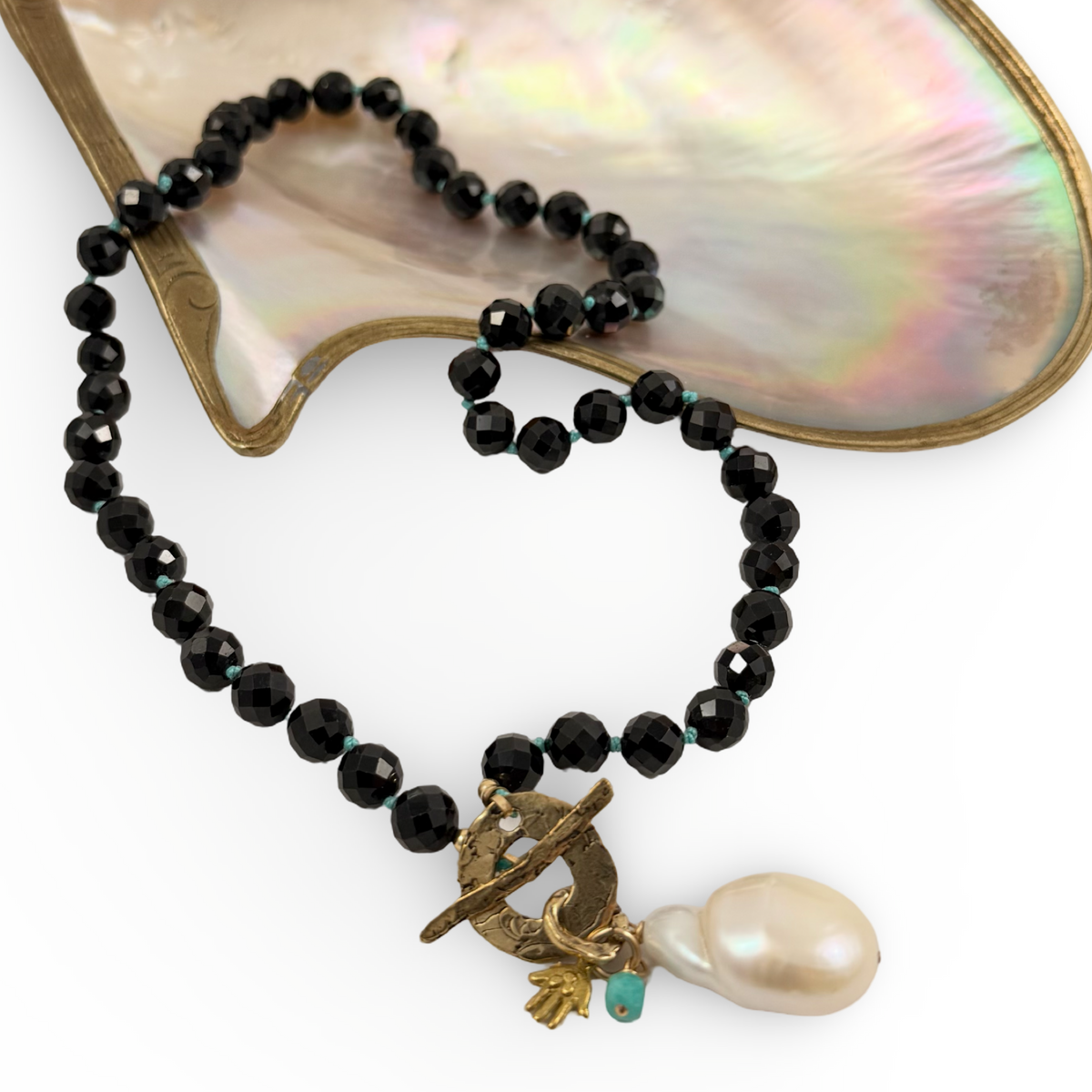 A beaded necklace with faceted black beads, a small turquoise accent, and a toggle clasp featuring a baroque pearl and gold-toned details, displayed on a white background with a mother-of-pearl shell prop.