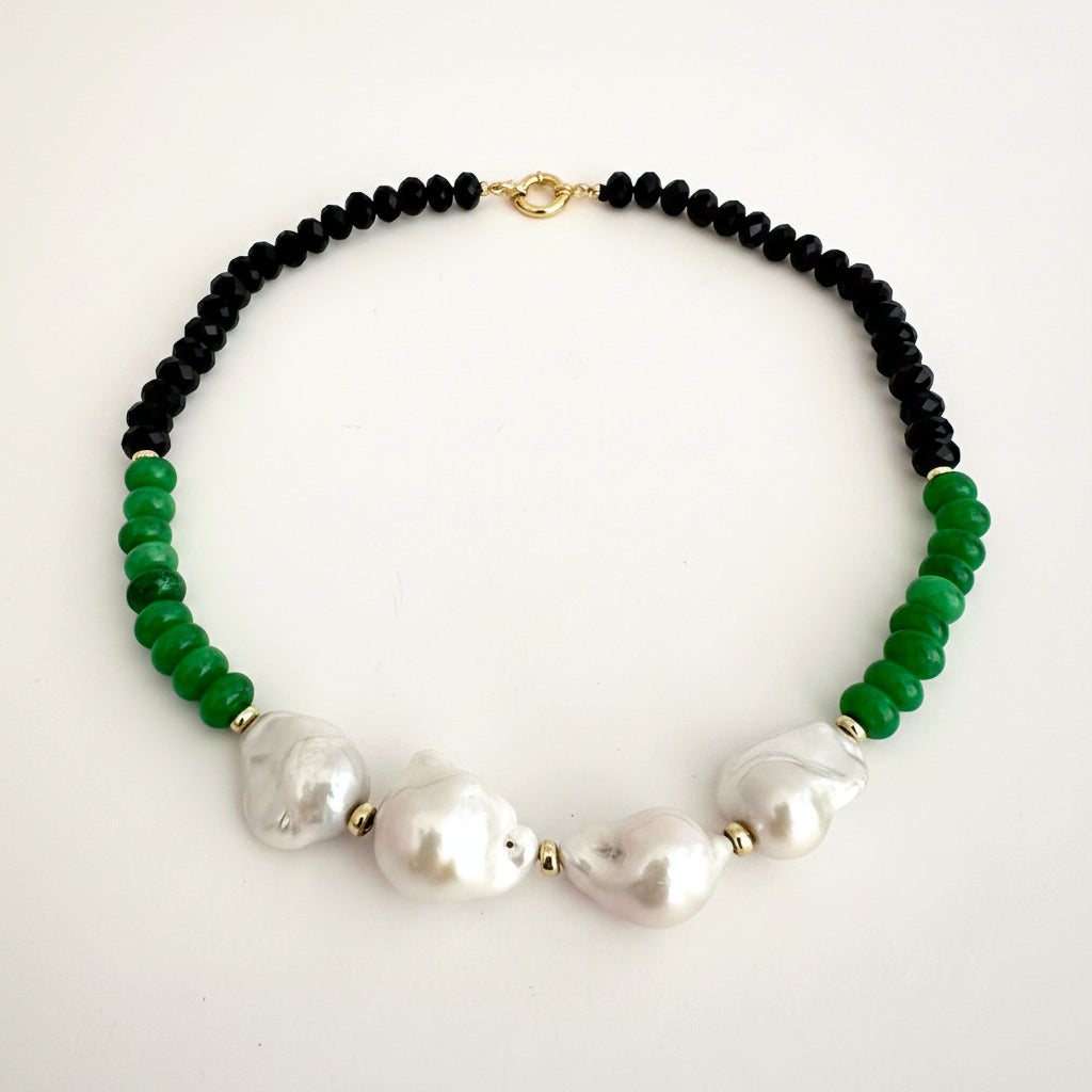 A beaded necklace featuring black round beads, vibrant green faceted beads, and three large white baroque pearls, accented with small gold spacers and a gold clasp, displayed on a clean white background.