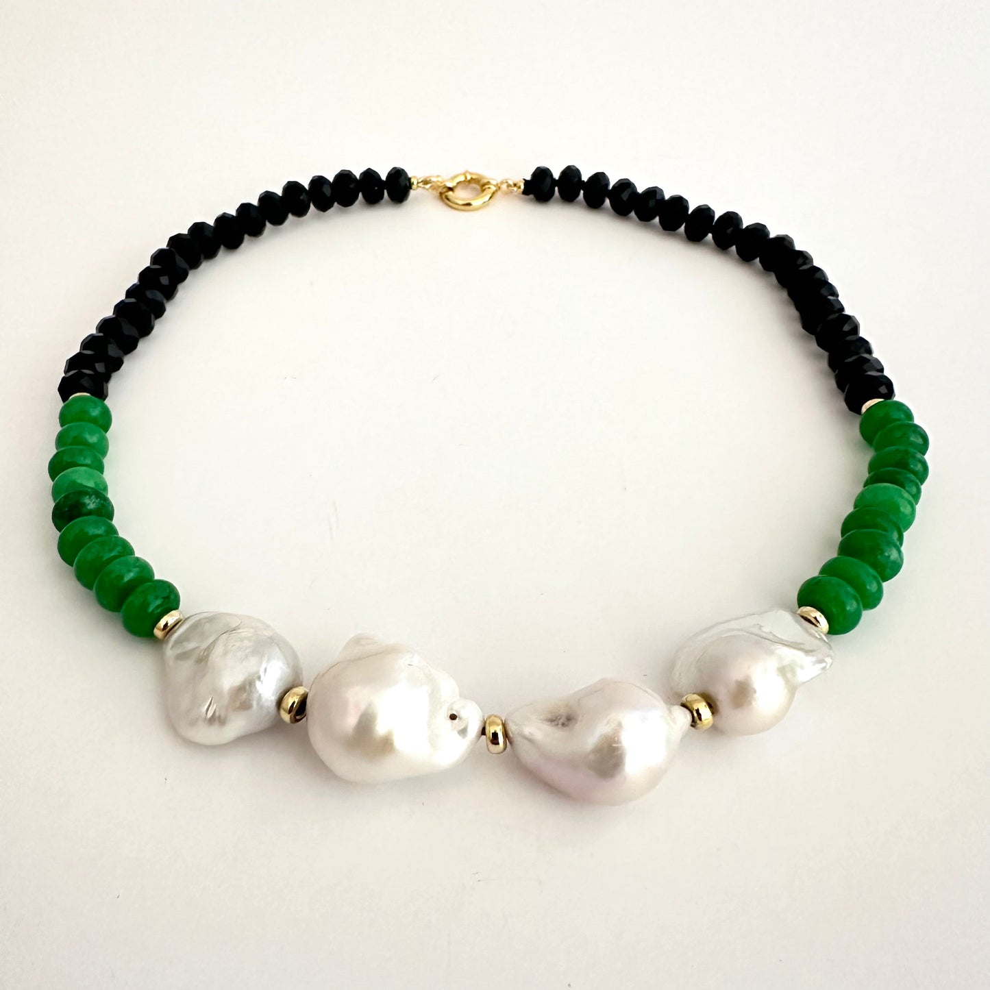 A beaded necklace featuring black round beads, vibrant green faceted beads, and four large white baroque pearls, accented with small gold spacers and a gold clasp, displayed on a clean white background.